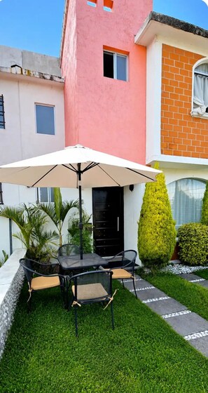 A patio with a table and chairs, surrounded by a well-maintained lawn and a building with a pink facade and a brick section. - House 5 minutes from the airport, Santa Fe, industrial area, covered porch, barbecue, and pet. (Lerma de Villada)
