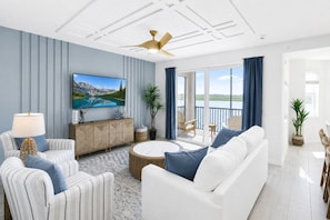 Ein modernes Wohnzimmer mit einem Flachbildfernseher, einem weißen Sofa, gestreiften Sesseln, einem runden Couchtisch und einem Balkon mit Seeblick.