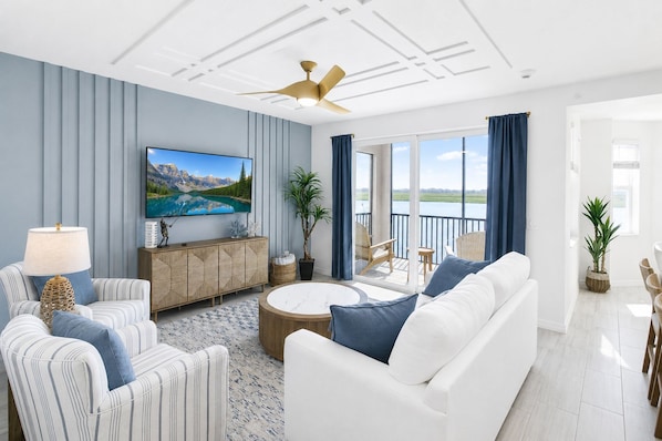 Ein modernes Wohnzimmer mit einem Flachbildfernseher, einem weißen Sofa, gestreiften Sesseln, einem runden Couchtisch und einem Balkon mit Seeblick.
