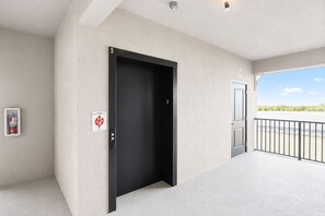 Ein moderner Gebäudeeingang mit einer schwarzen Tür, einem Feuerlöscher und freiem Blick auf Himmel und Grünflächen.