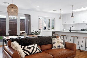 Eine moderne Küche mit weißen Schränken, ein Essbereich mit Tisch und Stühlen sowie ein Wohnzimmer mit braunem Ledersofa und dekorativen Kissen.