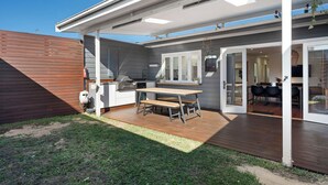 A covered outdoor patio with a wooden table and chairs, a barbecue grill, and a sliding glass door leading to an interior dining area. - Pet-friendly cottage near cafes, pubs and parks (Merewether)