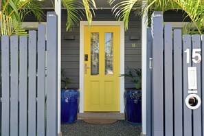 Une porte jaune, sur laquelle est inscrit le numéro « 15 », est flanquée de deux jardinières bleues et mène à une porte grise portant également le numéro « 15 », mais cette fois à droite.