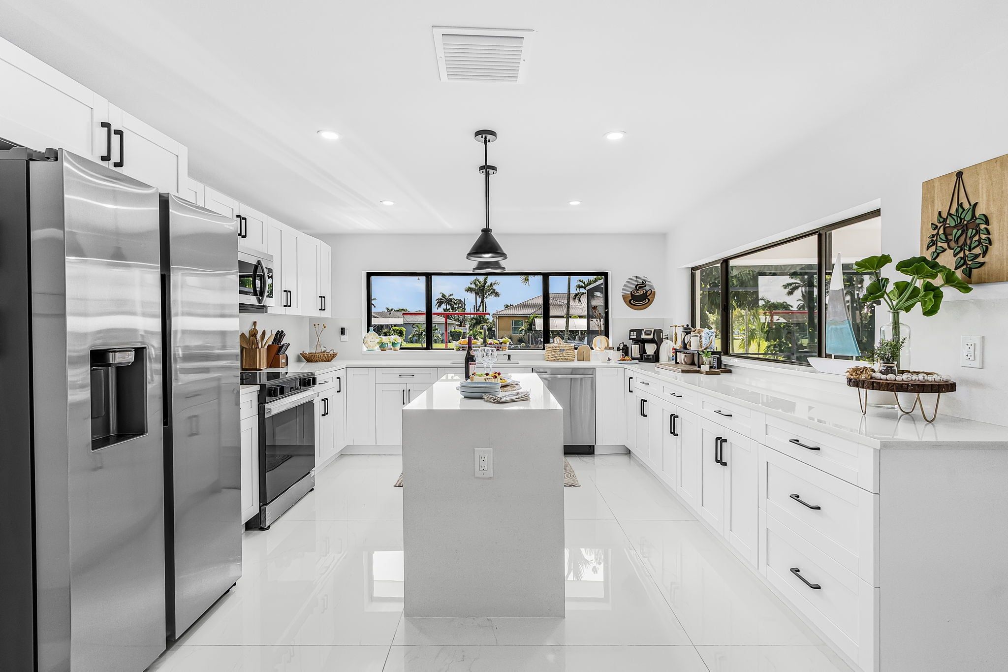 A modern kitchen with white cabinetry, stainless steel appliances, a central island, and a large window offering a view of palm trees.