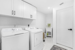 A laundry room with two white washing machines, a small table with a plant, and a door. - Gulf Access Getaway with Heated Pool, Dock, Kayaks and Fun Family Retreat - Salty Serenity - Roelens (Cape Coral)
