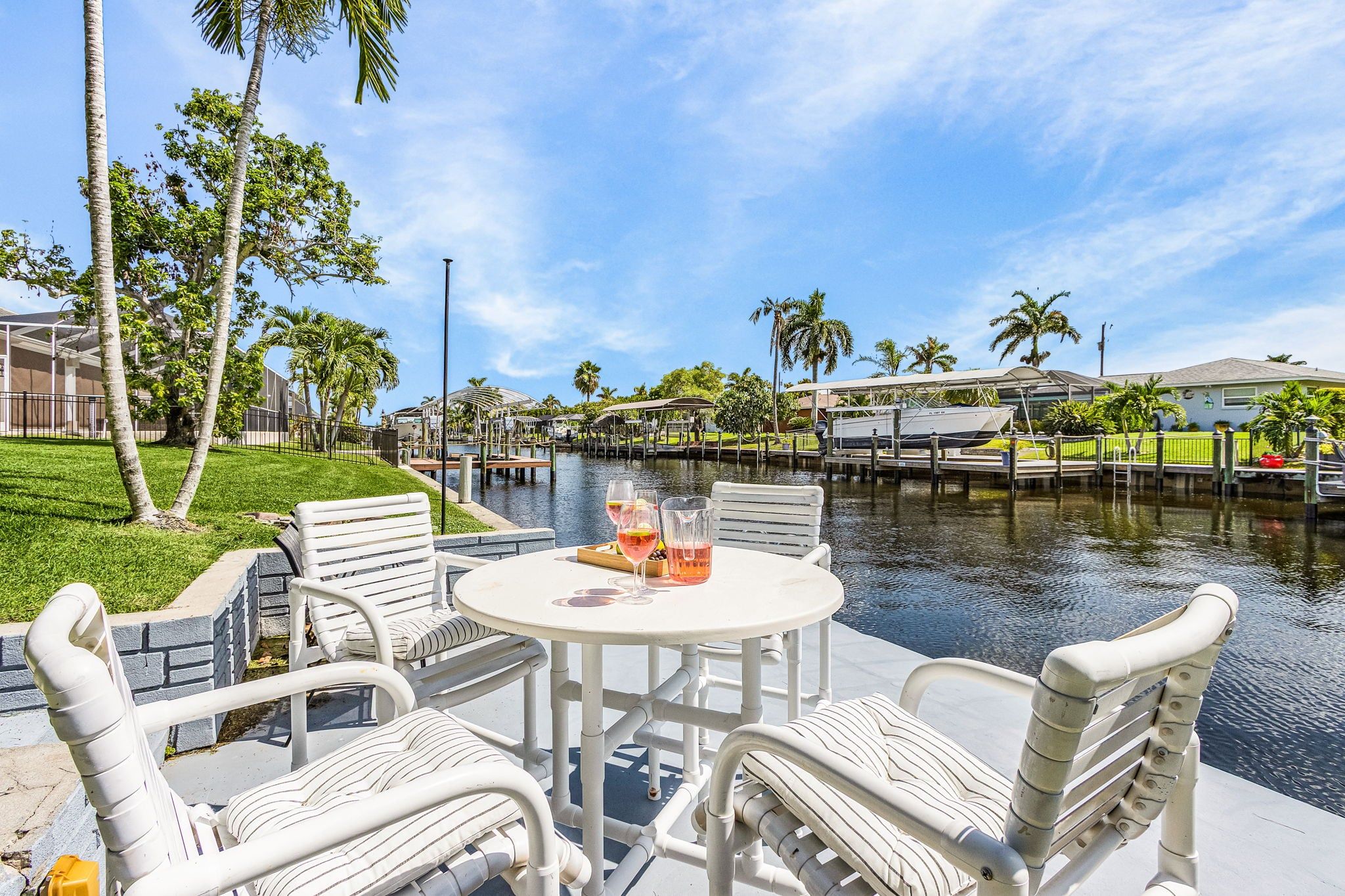 A waterfront patio with white wicker furniture, a table with drinks, and a view of the water and buildings in the background.