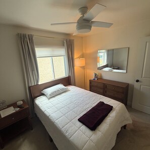 Ein Hotelzimmer mit Bett, Nachttisch, Lampe, Deckenventilator, Fenster mit Vorhängen und Tür.