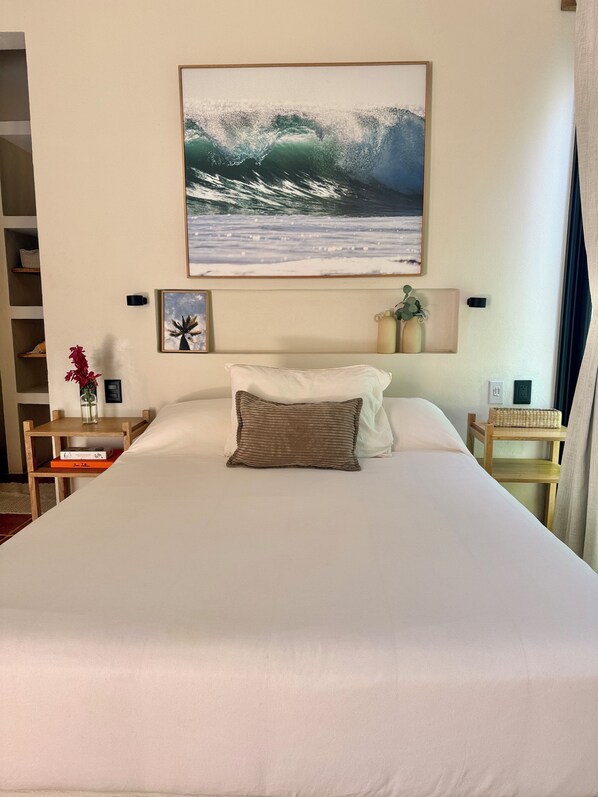 A neatly made bed with a pillow, a framed picture of a wave on the wall, and a small table with a vase and a book.