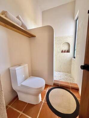 A bathroom with a toilet, a shower area with a tiled floor, and a small shelf with bottles.