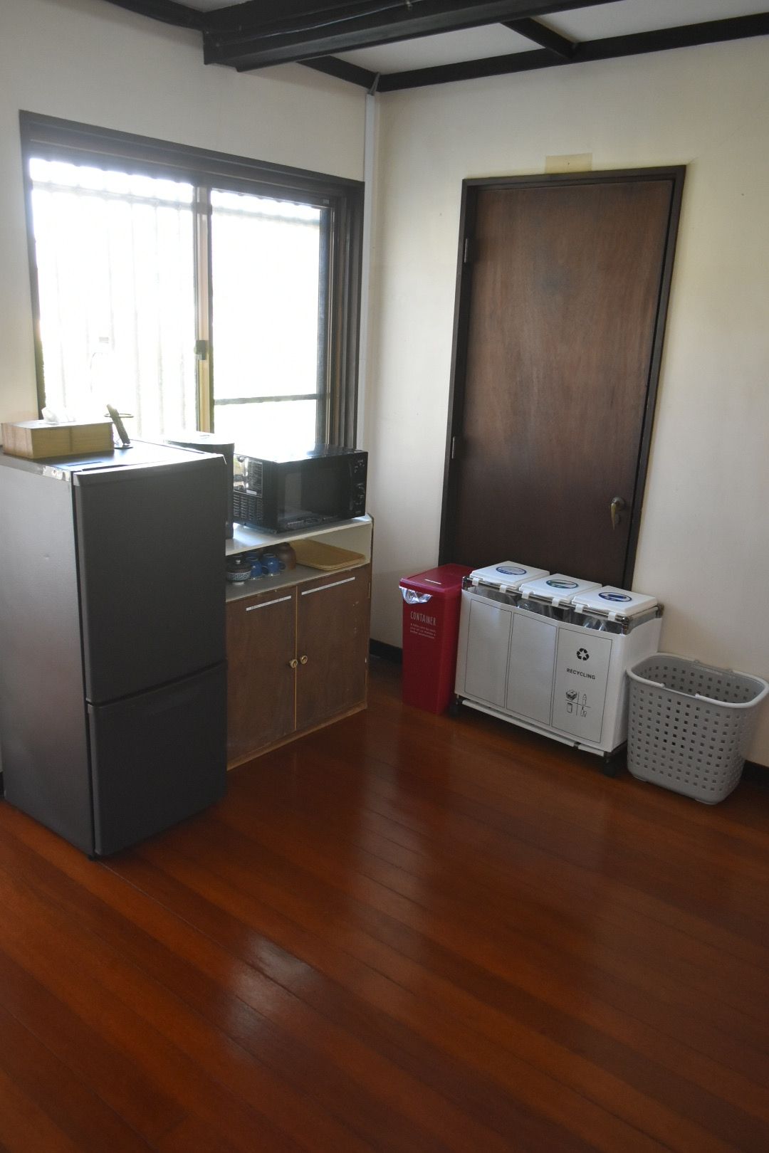 A compact kitchen with a refrigerator, microwave, and a small sink. There's a door and a trash bin visible.