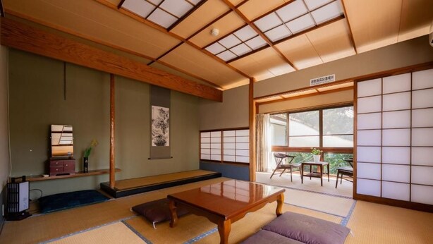 Una habitación japonesa tradicional con suelo de tatami, una mesa de madera baja, cojines, una puerta corredera y vistas al exterior.