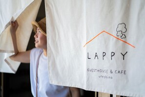 Una persona sostiene un paño blanco con el texto 'LAPPY GUESTHOUSE & CAFE Ureshino' y un sencillo icono de una casa.