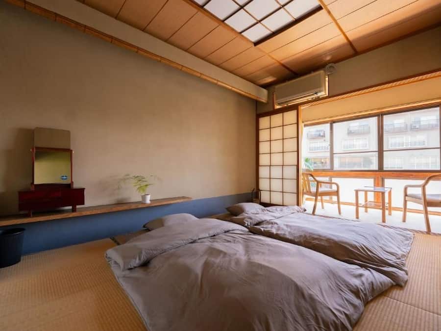 Quarto japonês tradicional com tatames, cama de madeira baixa, escrivaninha pequena com espelho e janela com vista para edifícios.