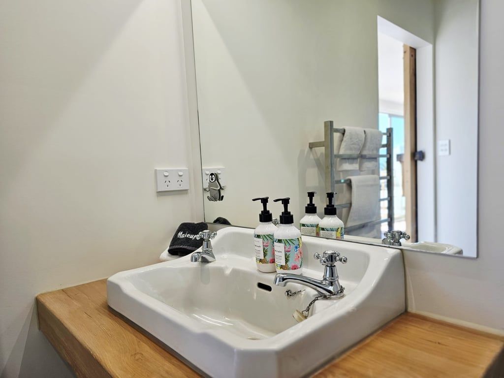 Une salle de bain avec un lavabo blanc, deux robinets et un miroir au-dessus. On y trouve trois flacons de savon liquide sur le lavabo.
