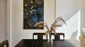 Une table à manger ornée d’un vase contenant des plantes séchées, un tableau accroché au mur et une fenêtre avec des stores.