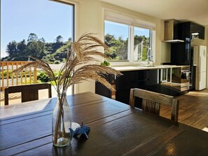 Una mesa de comedor con un jarrón de plantas secas, un juguete pequeño y vistas a una cocina y un espacio exterior.