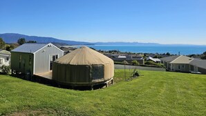 A yurt with a covered entrance, a white building with a sloped roof, and a view of the ocean and mountains in the background.