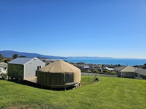 Una yurta con entrada cubierta, un edificio blanco con techo inclinado y una vista del océano y las montañas al fondo.