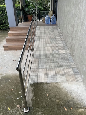 Een helling die leidt naar de ingang van een gebouw, met voelbare tegels voor blinden, een metalen leuning en een betonnen vloer.