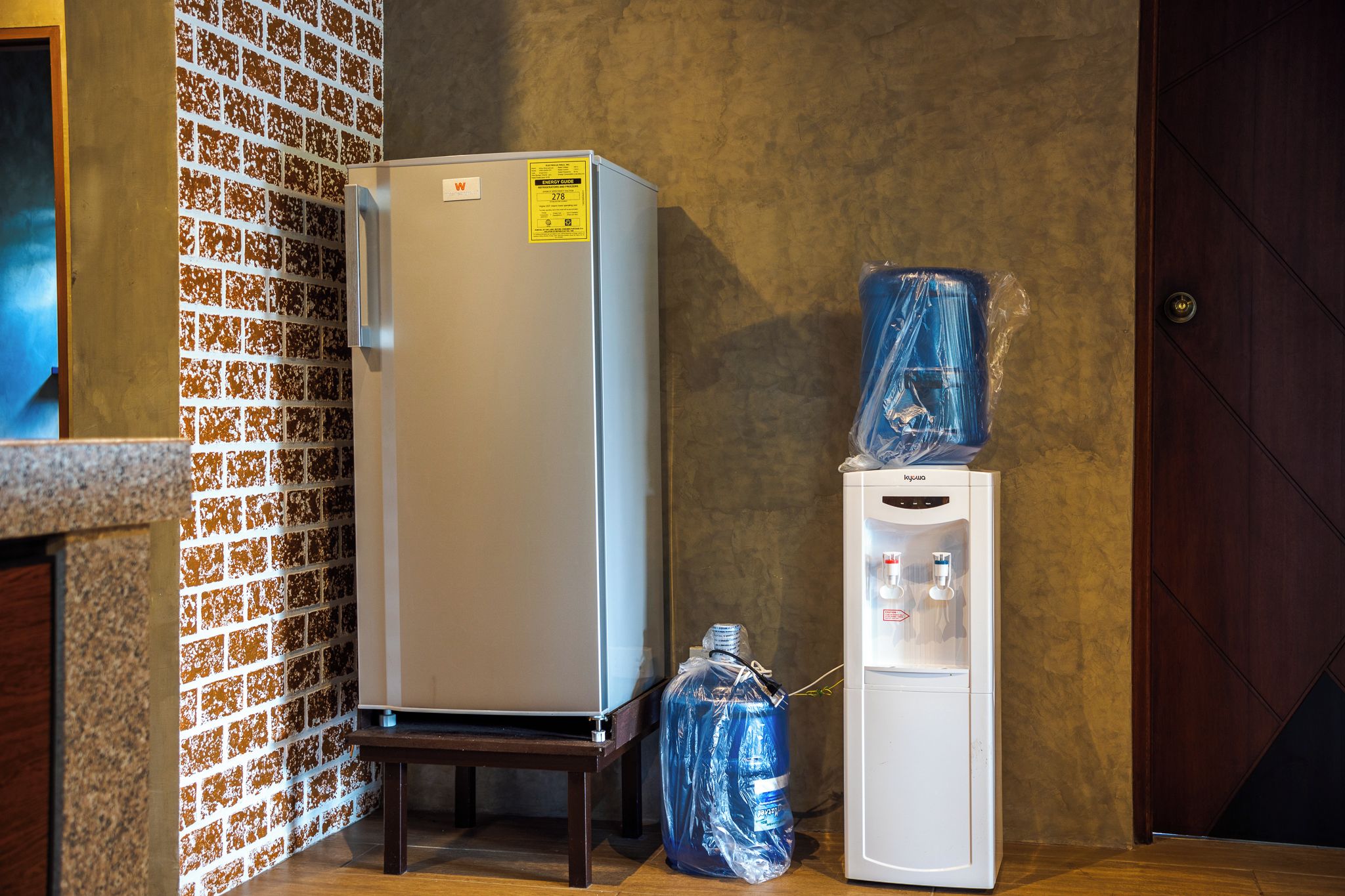 Ein Kühlschrank und ein Wasserspender in einem Raum mit einer Ziegelwand und einer Holztür.