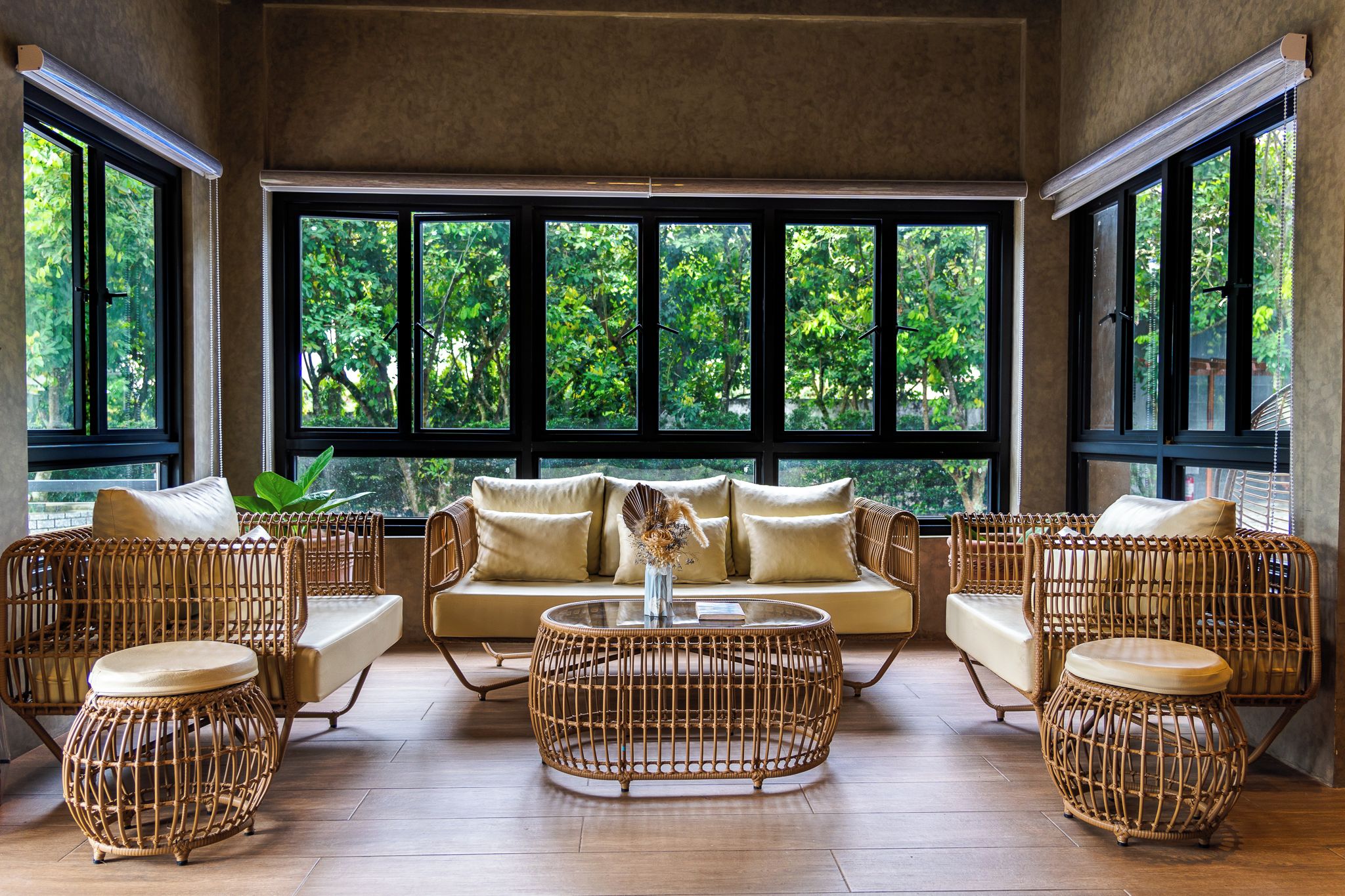 A room with a sofa set, two round wicker coffee tables, and large windows with a view of greenery.
