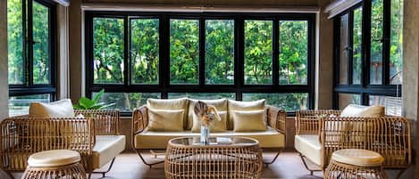 A room with a sofa set, two round wicker coffee tables, and large windows with a view of greenery.