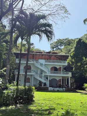 A two-story building with a white exterior and a red-tiled roof, surrounded by lush greenery and palm trees.