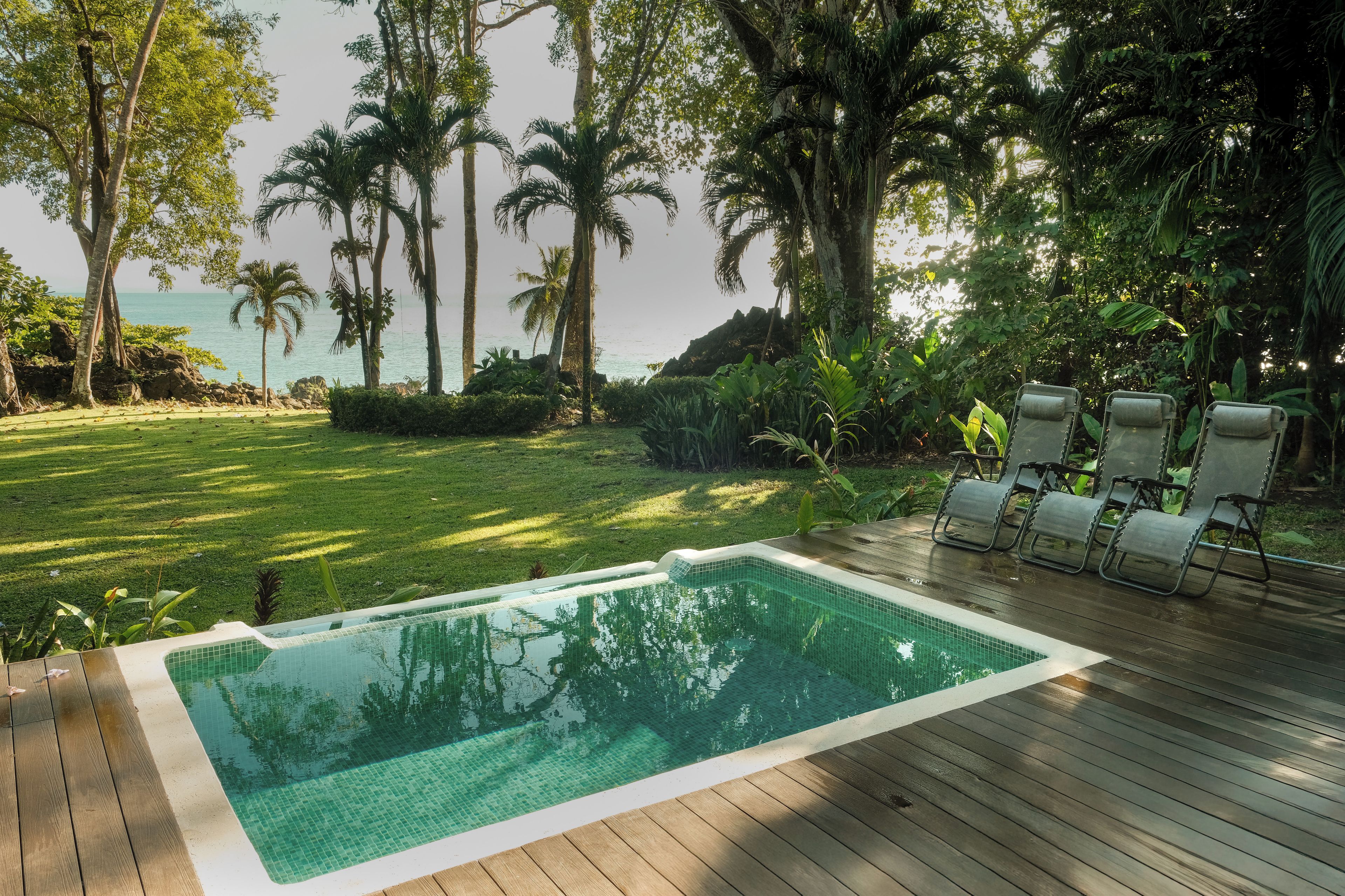 A swimming pool with a wooden deck, surrounded by lush greenery and palm trees, overlooking a body of water.
