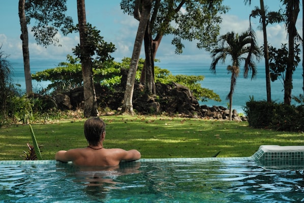 Una persona relajándose en una piscina con vista al océano y árboles tropicales.