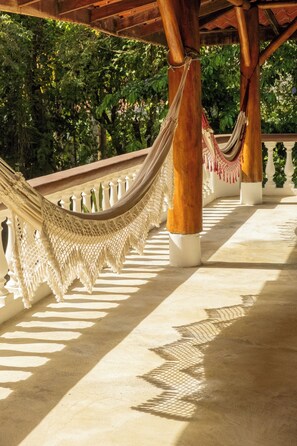 A hammock under a wooden structure with a white balustrade and greenery in the background.