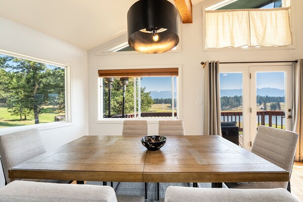 Un coin repas avec une grande table en bois, des chaises et un saladier. Des fenêtres offrent une vue sur le paysage.