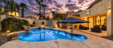 A swimming pool with a patio area, surrounded by a landscaped garden and a house with a tiled roof.