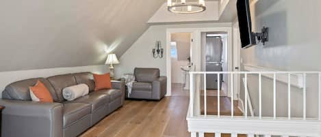 A spacious living room with a grey leather sofa, a white staircase, and a flat-screen TV mounted on the wall.