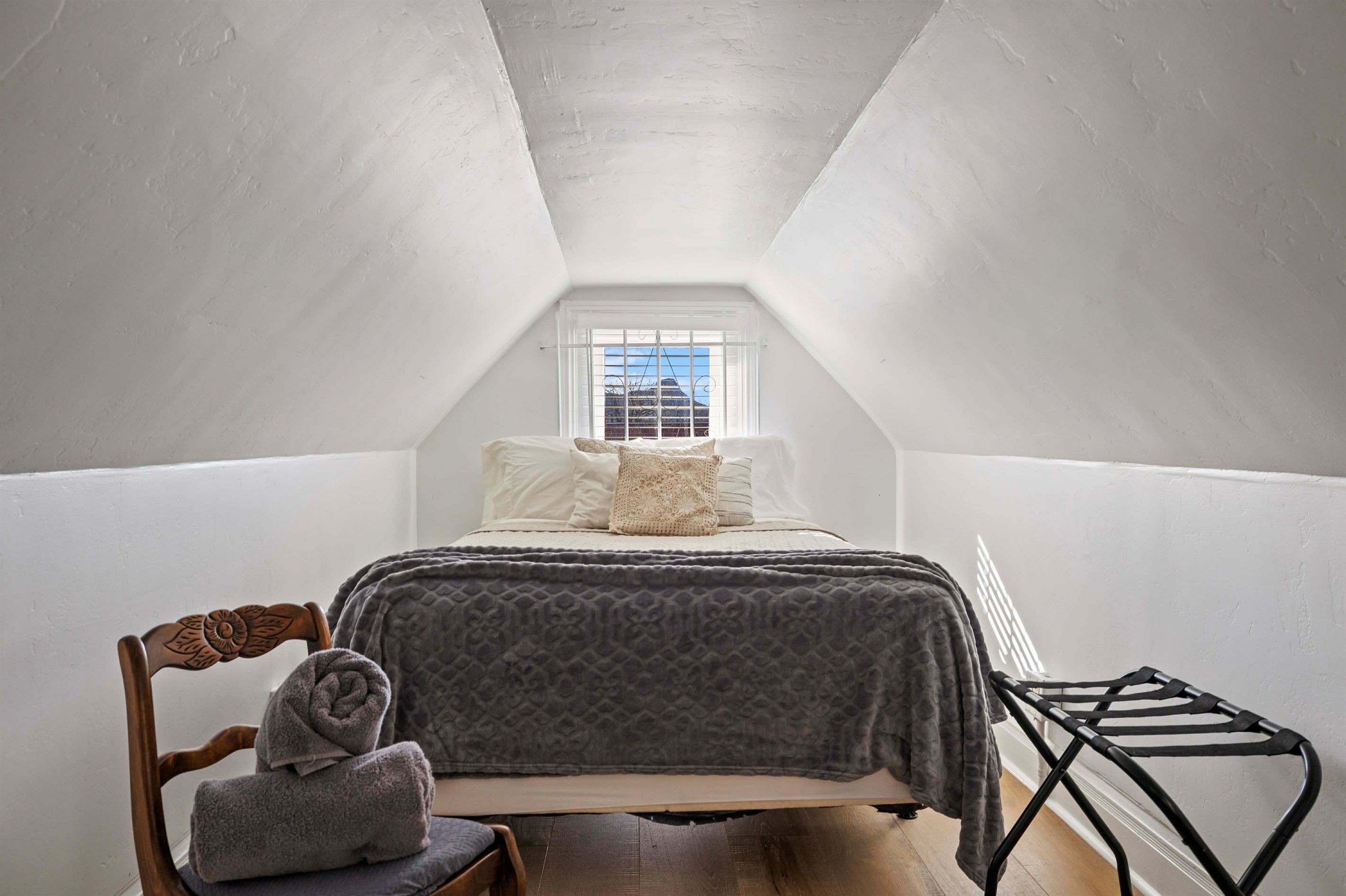 Un dormitorio abuhardillado pequeño y triangular con una cama, una silla y un ventanal.