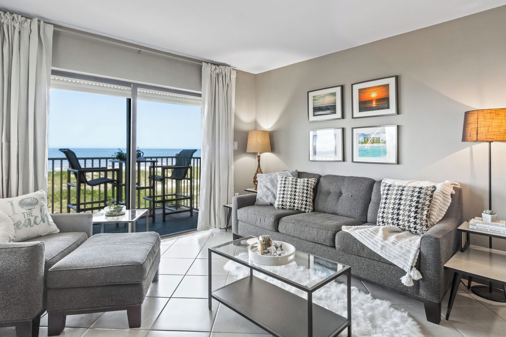 A modern living room with a grey sofa, a glass coffee table, and a view of the sea through large windows.