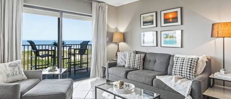 A modern living room with a grey sofa, a glass coffee table, and a view of the sea through large windows.