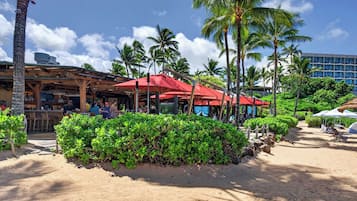 A beachfront bar area with red umbrellas, palm trees, and a sandy path.