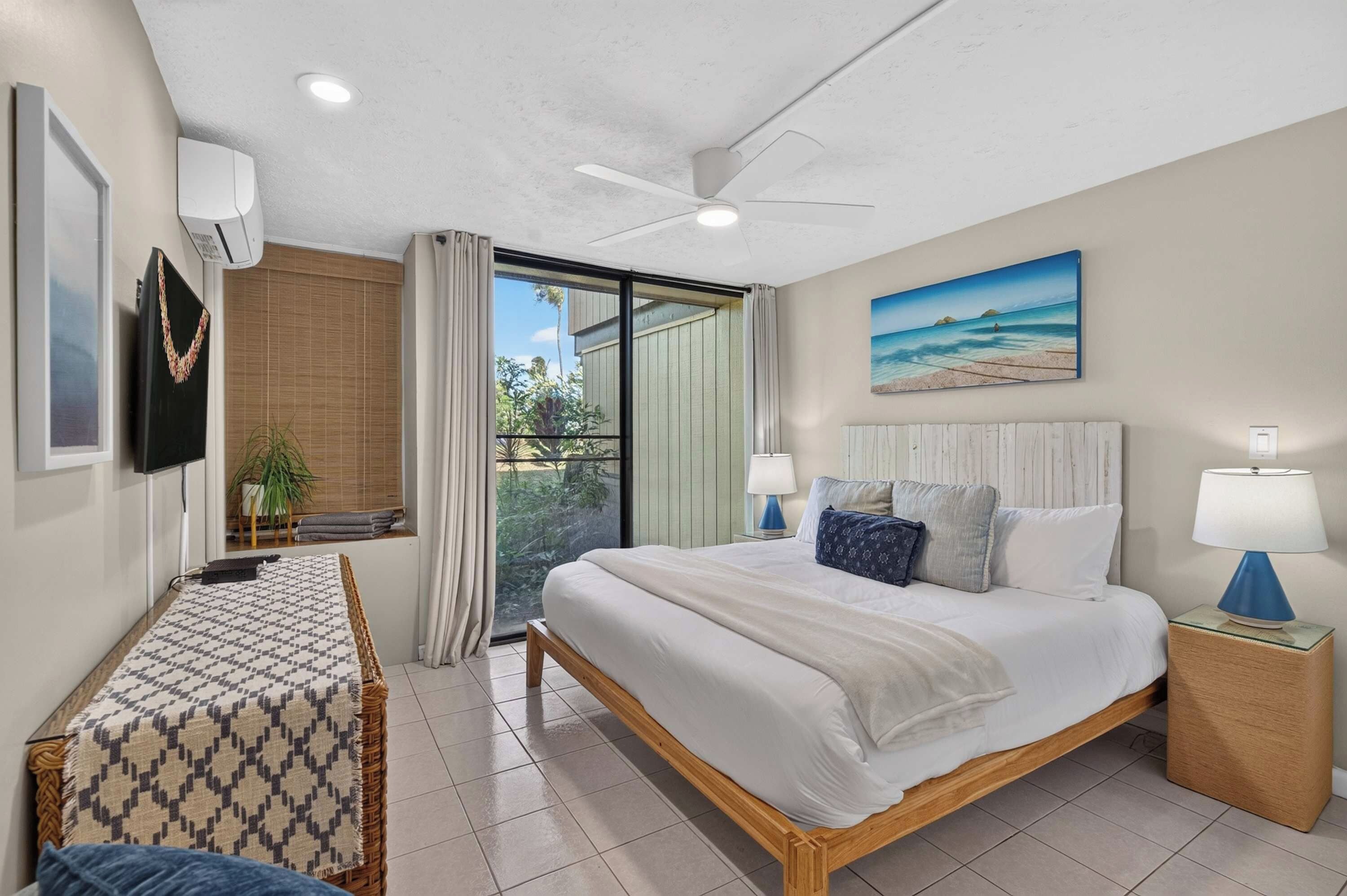 A hotel room with a bed, a desk, a ceiling fan, a window with a view, and a painting on the wall.