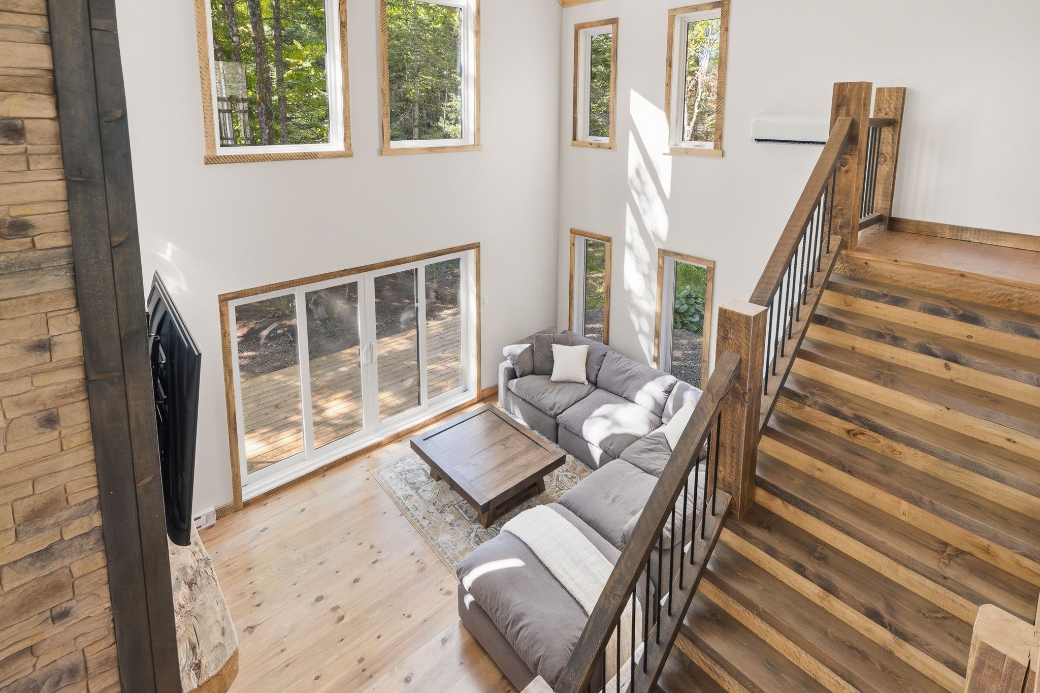 A spacious living area with a wooden staircase, a grey sofa set, and large windows offering views of the outdoors.