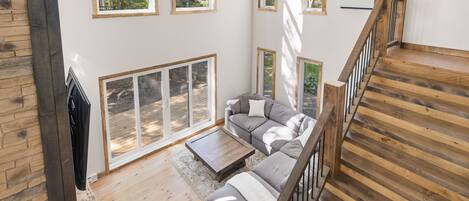 A spacious living area with a wooden staircase, a grey sofa set, and large windows offering views of the outdoors.