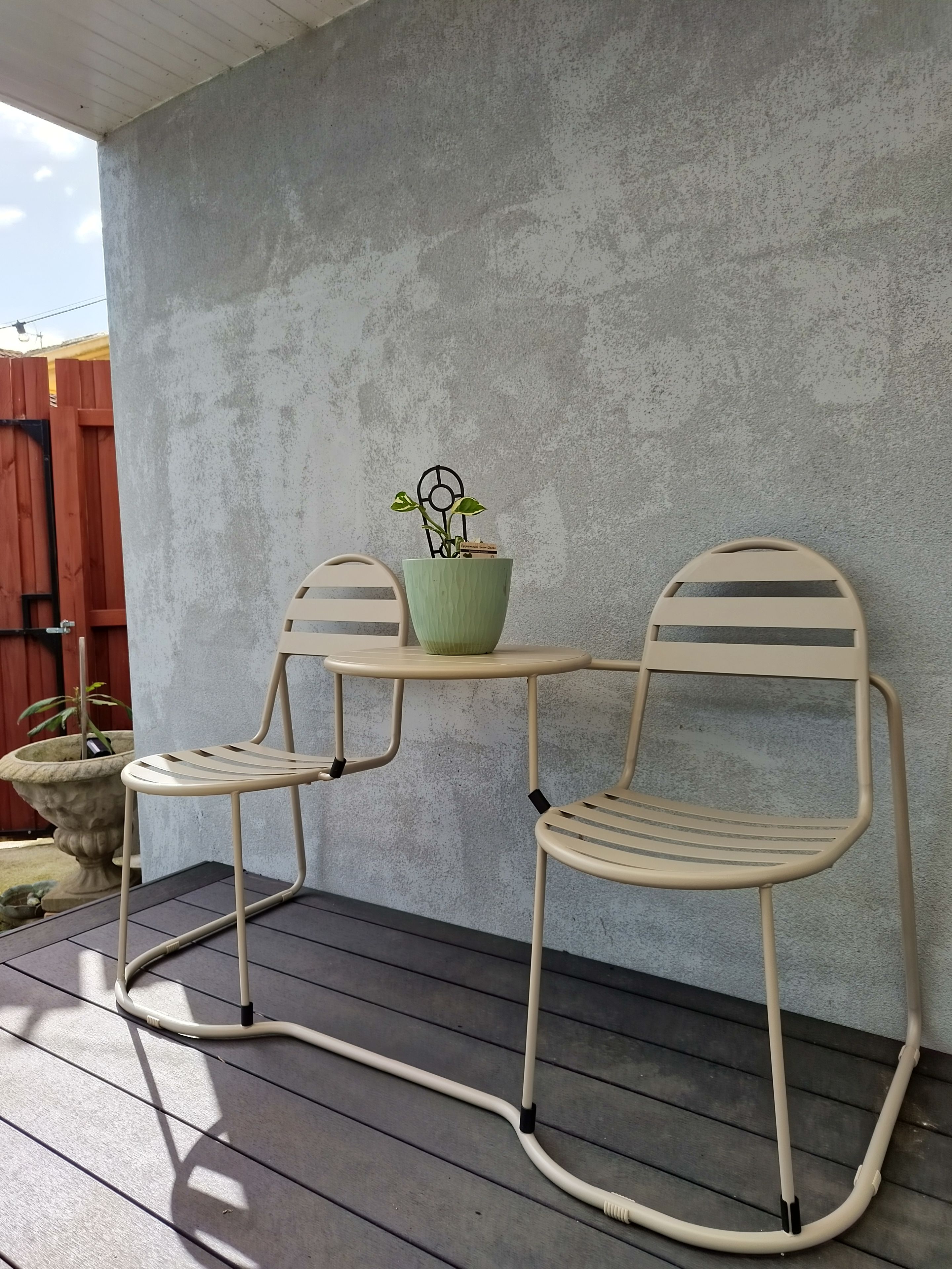 Een terras met twee metalen stoelen en een tafel, een potplant en een betonnen muur.