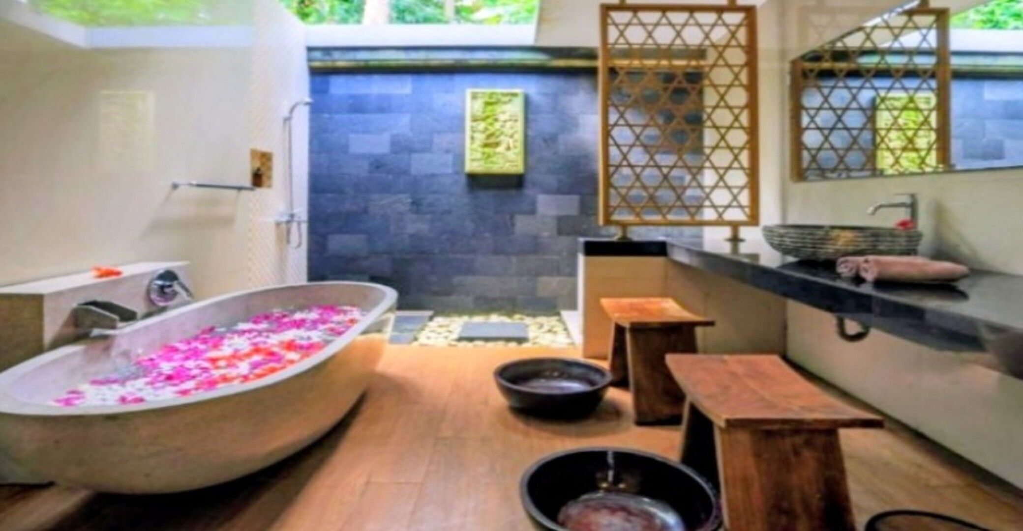 A bathroom with a freestanding bathtub filled with rose petals, a wooden bench, and a stone wall.