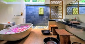 A bathroom with a freestanding bathtub filled with rose petals, a wooden bench, and a stone wall in the background. - Cozzy Fullmoon Purnama Villa Ubud (Gianyar)