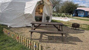 A white tent with an open entrance, a wooden picnic table, and a small black stove. - The Leaf & Lantern Dome - Sleeps 5 - Dog Friendly (Pewsey)