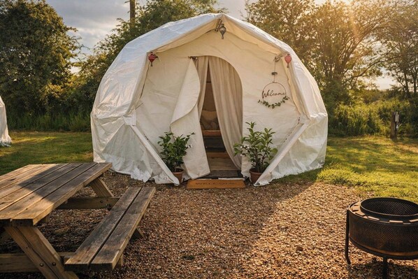 Una tenda amb una porta, dues plantes en testos, una banca de fusta i una barbacoa.
