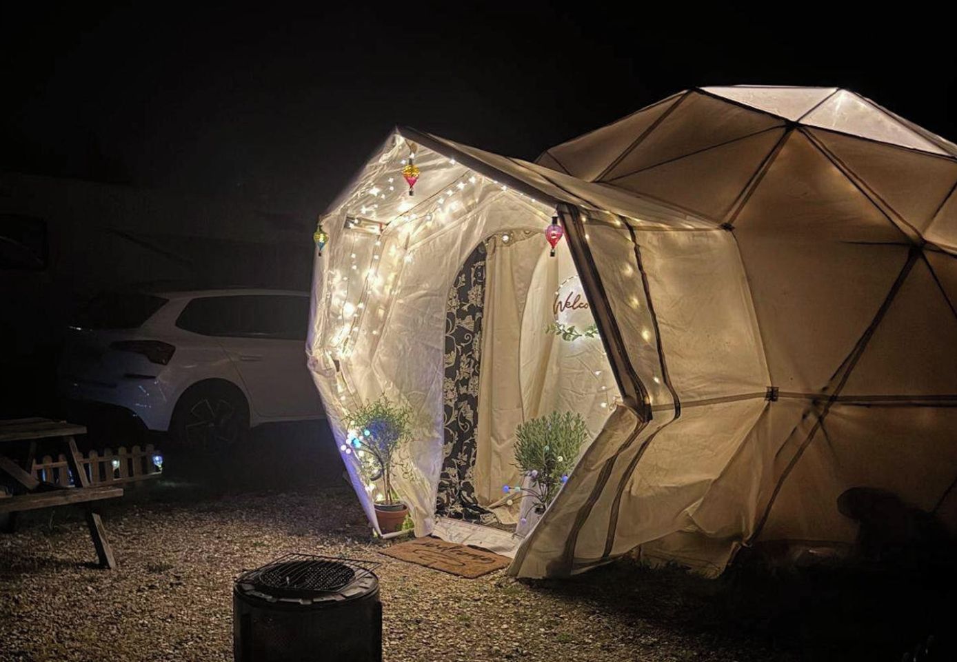 Een tent met een luifel en lichtjes, een geparkeerde auto in de buurt, en een klein tafeltje met spullen erop.