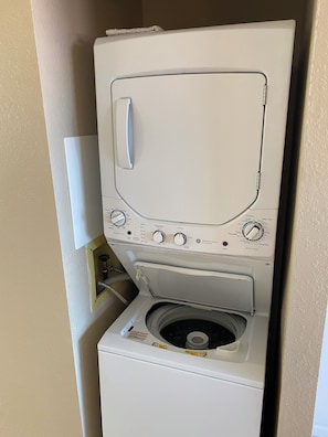 A front-loading washing machine with a control panel, a dryer below, and a wall-mounted electrical outlet. - Ocean View 2/2, 2 Cal-King Beds, Washer Dryer, Golf Clubs, Tennis Hot tub,Honalo (Holualoa)