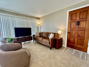 A living room with a brown sofa, a television, a lamp, and a wooden door. - Ocean View 2/2, 2 Cal-King Beds, Washer Dryer, Golf Clubs, Tennis Hot tub,Honalo (Holualoa)