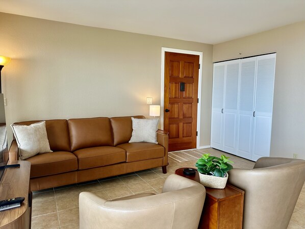 Una sala de estar con un sofá de cuero marrón, un sillón beige, una mesita de madera y una planta en maceta.