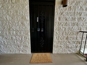 A black door with a number plate, a textured white wall, and a doormat with a pineapple design. - Ocean View 2/2, 2 Cal-King Beds, Washer Dryer, Golf Clubs, Tennis Hot tub,Honalo (Holualoa)